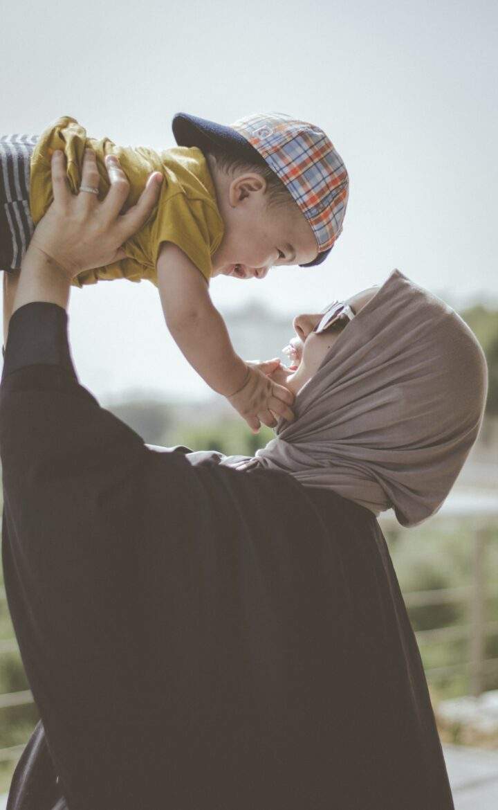 Mom with hijab and sunglasses lifts smiling baby with backwards cap into the air