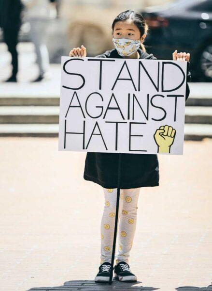 east-asian child protester wearing mask holding sign that says "stand against hate"