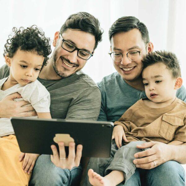 two dads and their young children all look at something on a tablet